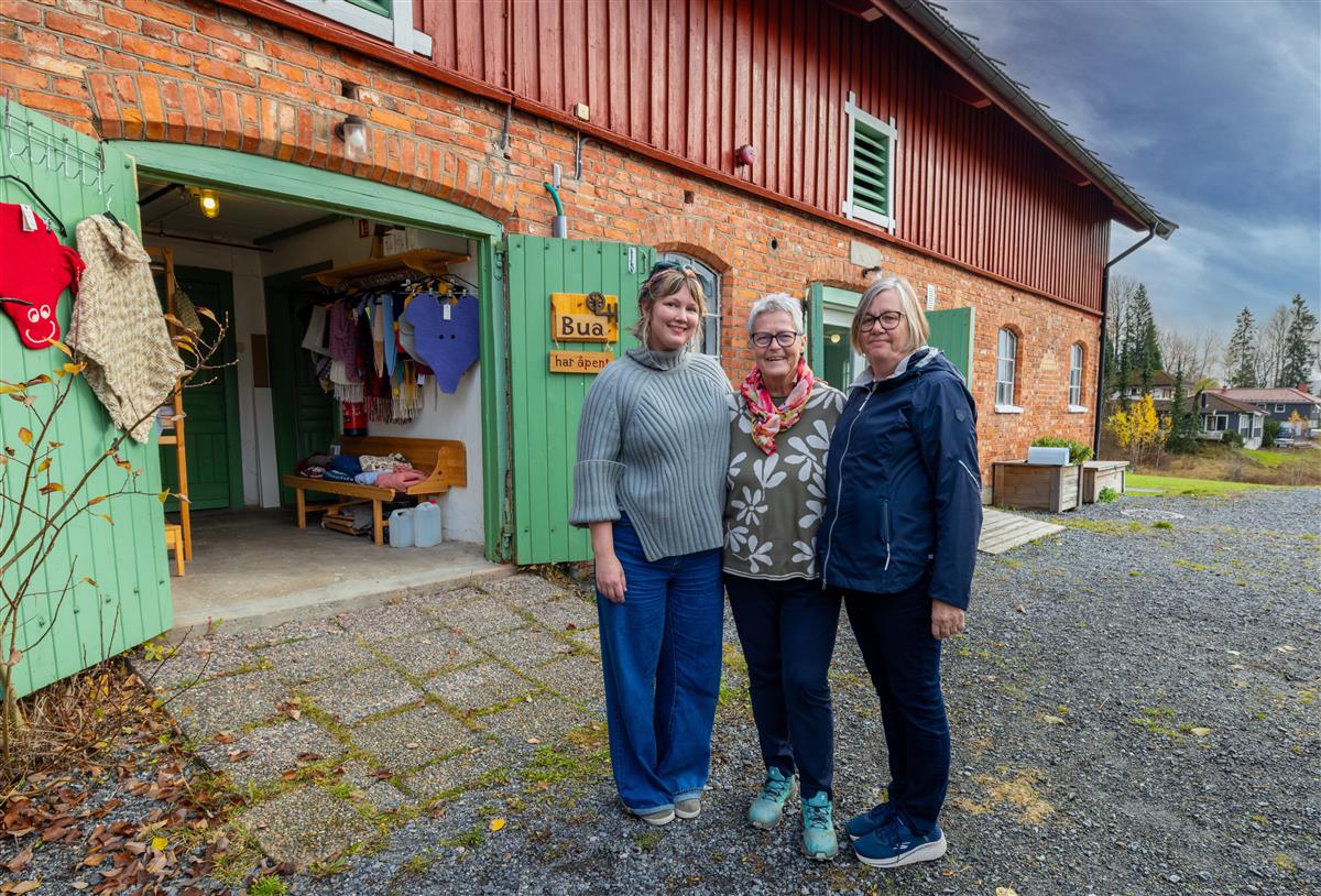 Tre kvinner står utenfor en rustikk murbygning med grønne dører og viser frem fargerike gjenstander til salgs. - Klikk for stort bilde
