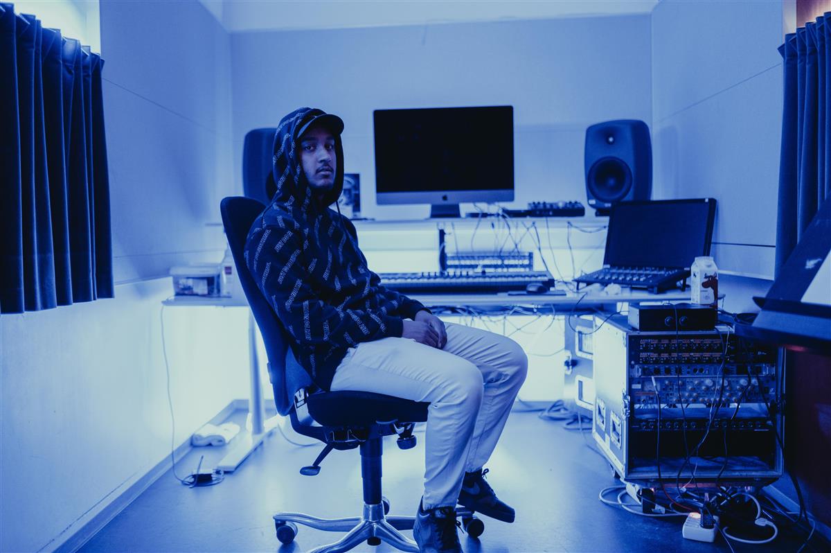 En ung mann i en kontorstol sitter i et musikkstudio med utstyr og PC bak seg - Klikk for stort bilde