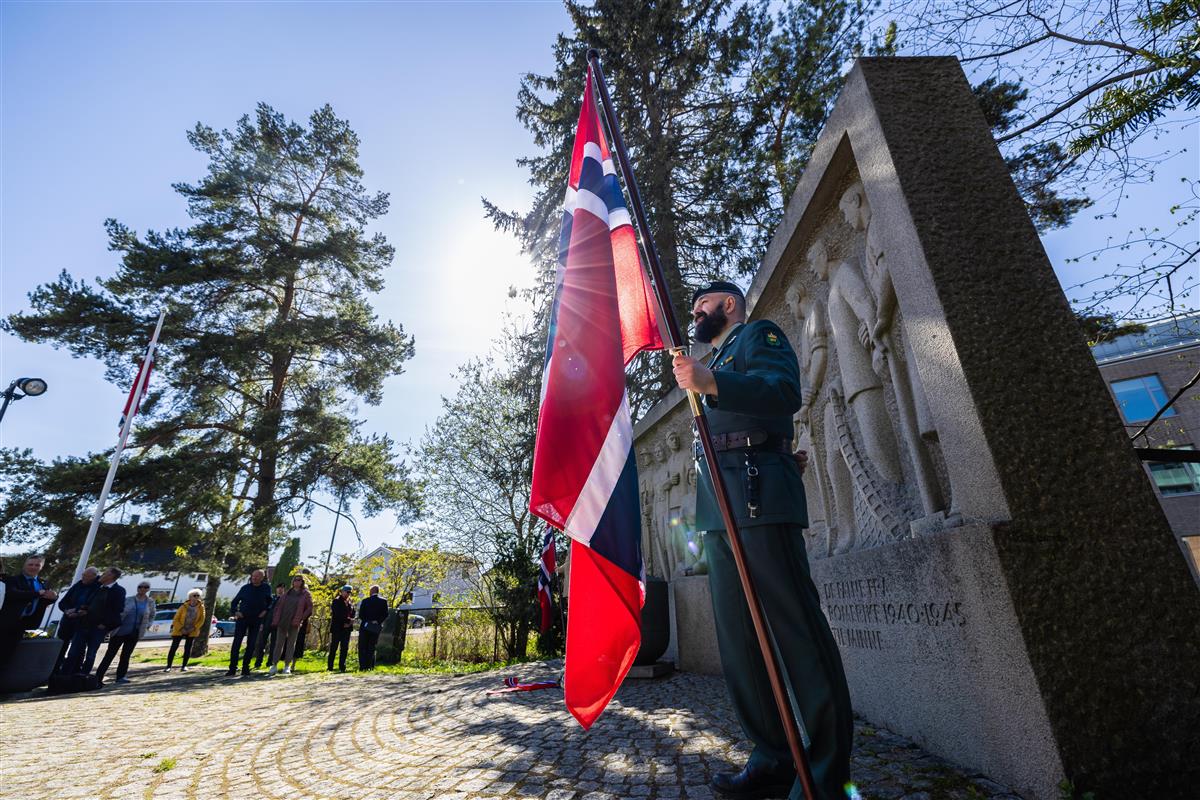 En person i uniform holder det norske flagget foran et monument med inskripsjoner. Solen skinner gjennom trærne i bakgrunnen, og flere mennesker kan ses rundt monumentet. - Klikk for stort bilde